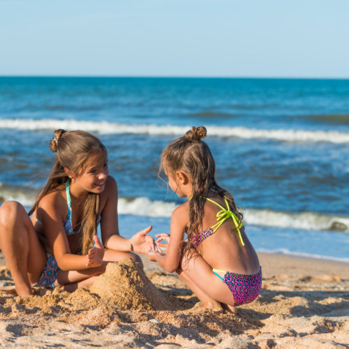 Two positive little girls sisters are building a sand castle sitting on the seashore on a sunny warm summer day. Concept of active children's games.